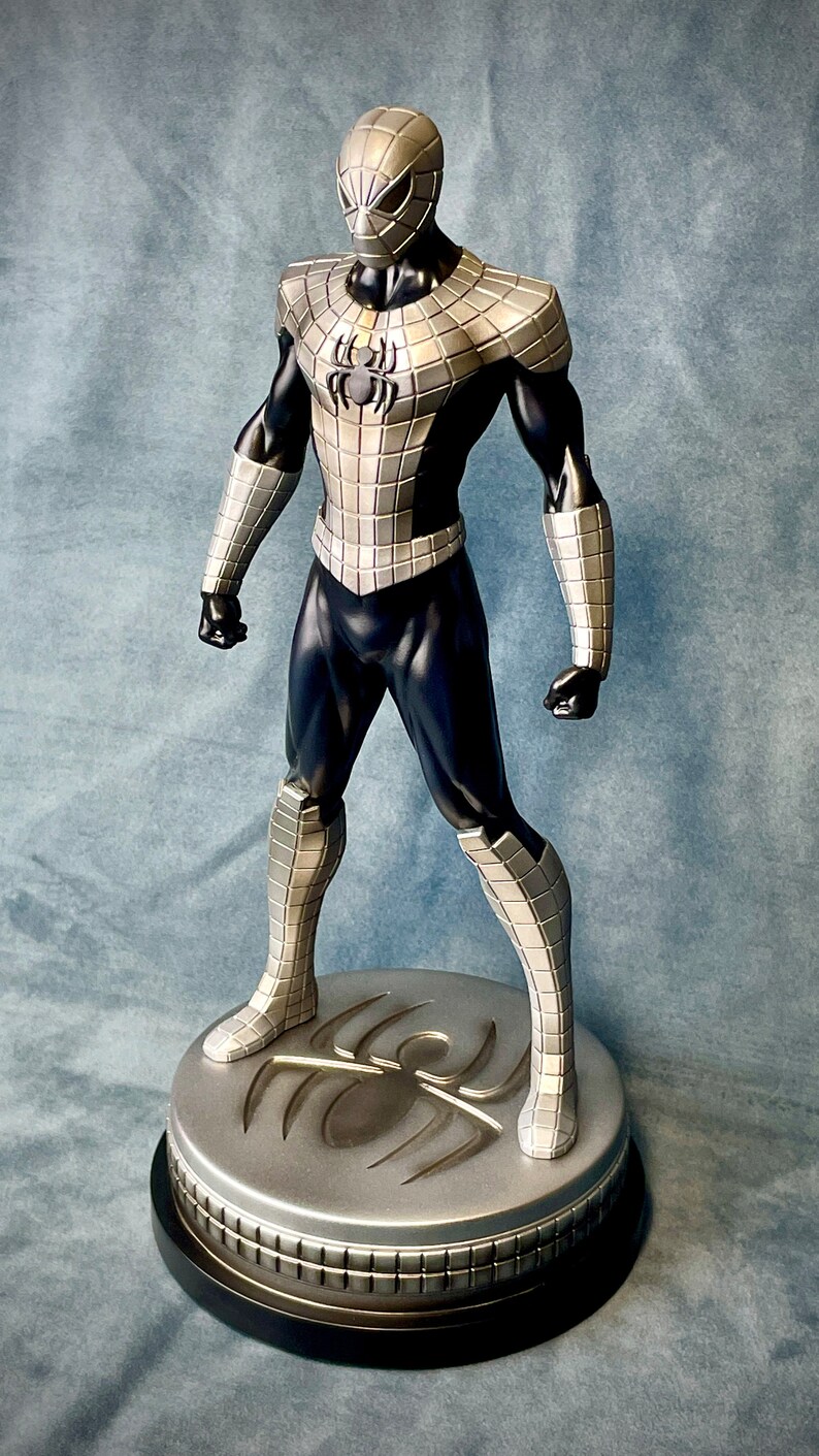 May include: A silver and black action figure of Spider-Man in a metallic suit. The figure is standing on a circular base with a spider logo.
