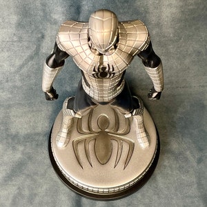 May include: A silver and black action figure of Spider-Man in a silver and black suit standing on a round base with a silver spider logo.