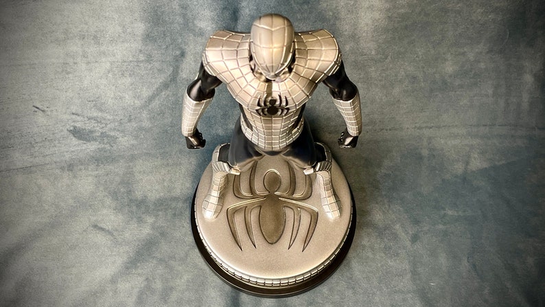 May include: A silver and black action figure of Spider-Man in a silver and black suit standing on a round base with a silver spider logo.