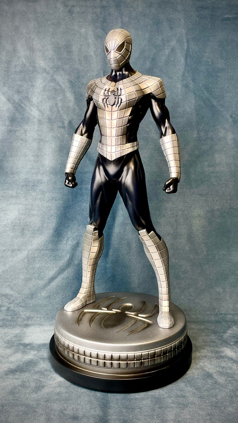 May include: A silver and black action figure of Spider-Man standing on a circular base with a spider logo. The figure is wearing a silver and black suit with a spider logo on the chest.