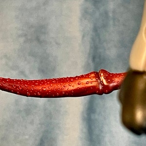 May include: A red, curved, plastic blade with water droplets on its surface. The blade is part of a toy action figure.