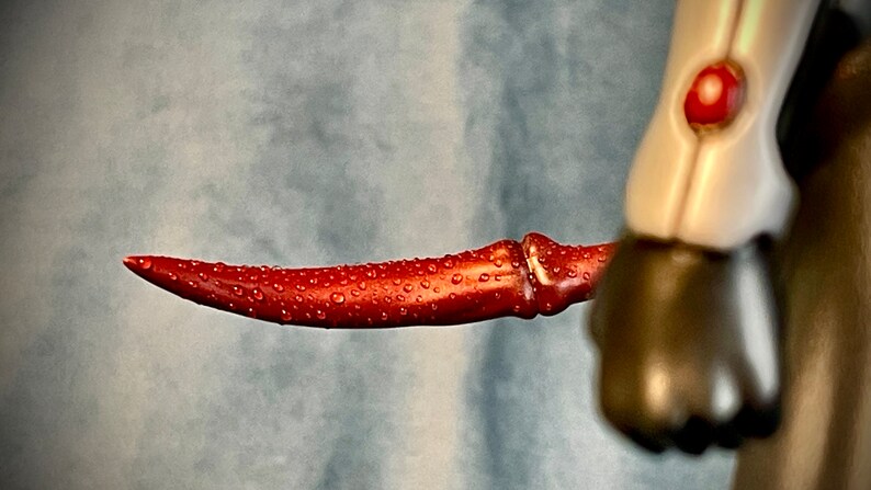 May include: A red, curved, plastic blade with water droplets on its surface. The blade is part of a toy action figure.