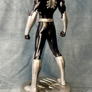 May include: A black and silver action figure of a superhero wearing a mask and a suit with a spider design on the back. The figure is standing on a silver base with a spider design.