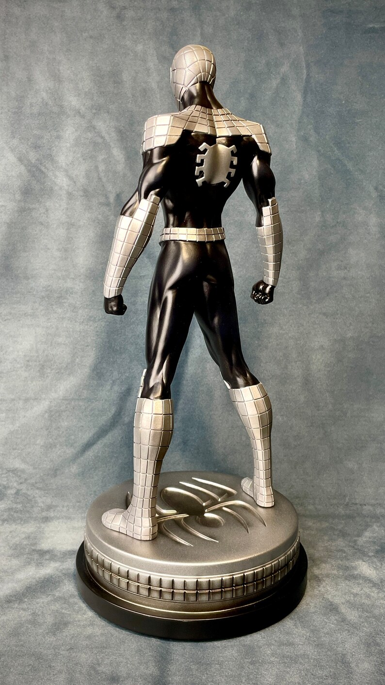 May include: A black and silver action figure of a superhero wearing a mask and a suit with a spider design on the back. The figure is standing on a silver base with a spider design.