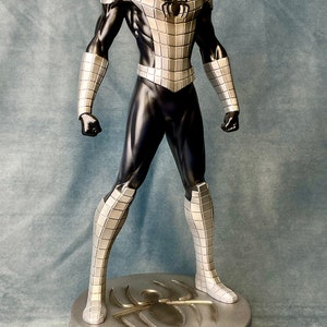May include: A silver and black action figure of Spider-Man standing on a silver base with a spider logo. The figure is wearing a silver and black suit with a black mask.
