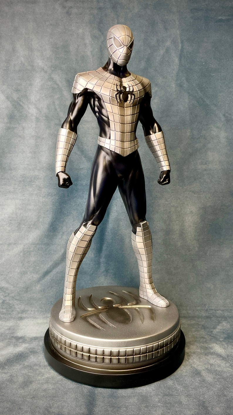 May include: A silver and black action figure of Spider-Man standing on a silver base with a spider logo. The figure is wearing a silver and black suit with a black mask.