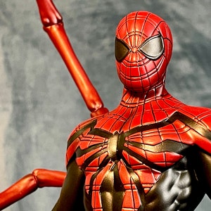 May include: A red and black Spider-Man action figure with a unique design. The figure has a red and black suit with a spider web pattern and a red mask with black eyes. The figure is posed with its arms outstretched and its legs slightly bent. The figure is standing on a gray background.