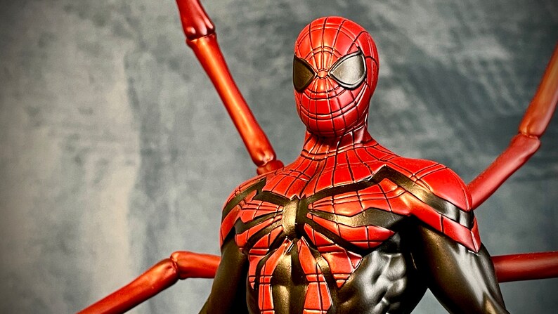 May include: A red and black Spider-Man action figure with a unique design. The figure has a red and black suit with a spider web pattern and a red mask with black eyes. The figure is posed with its arms outstretched and its legs slightly bent. The figure is standing on a gray background.