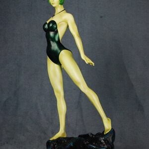 May include: A green-skinned figurine of a woman wearing a dark green swimsuit. The figurine is standing on a rocky base.