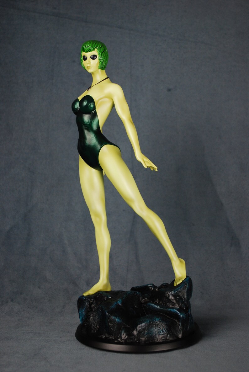 May include: A green-skinned figurine of a woman wearing a dark green swimsuit. The figurine is standing on a rocky base.