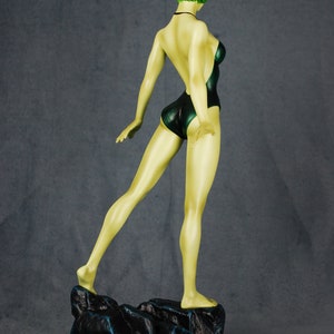 May include: A green-haired figurine of a person wearing a dark green swimsuit standing on a black rock base.