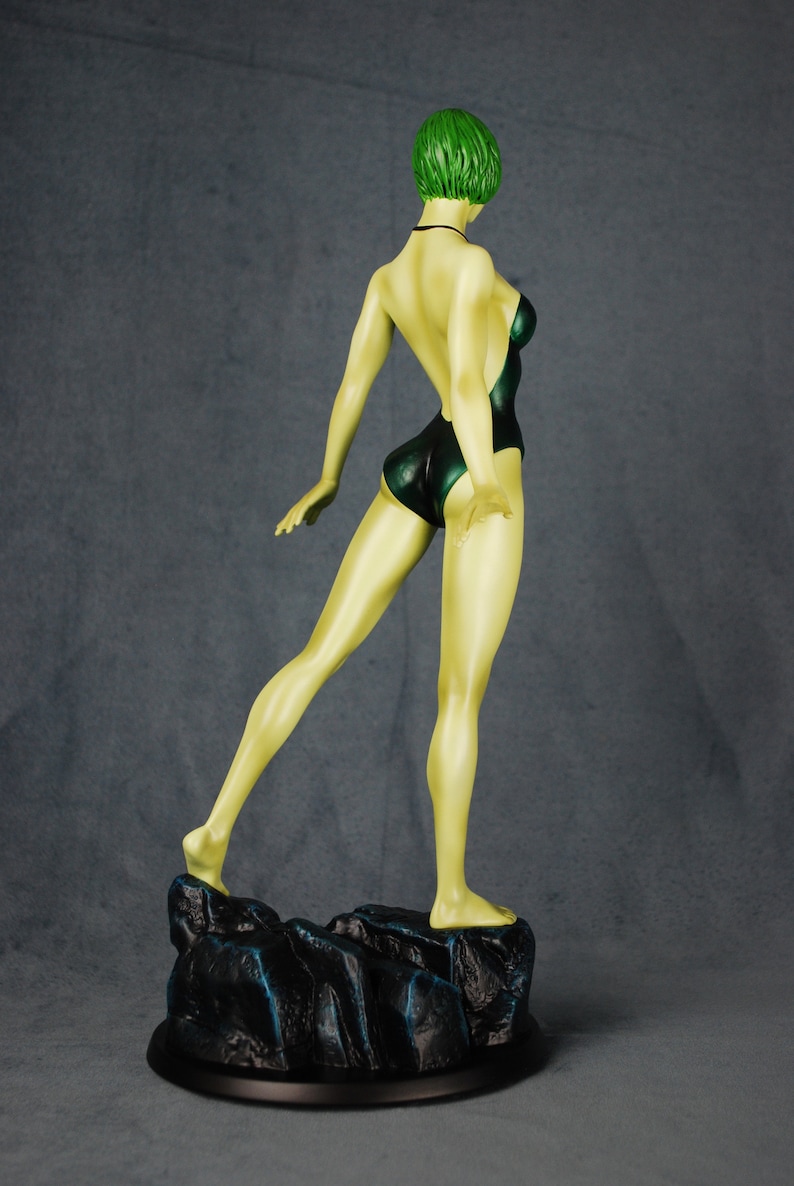 May include: A green-haired figurine of a person wearing a dark green swimsuit standing on a black rock base.