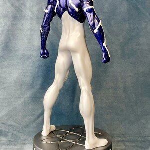 Puede incluir: Una figura de acci&oacute;n de un superh&eacute;roe de color blanco y azul de pie sobre una base plateada con un dise&ntilde;o de s&iacute;mbolo at&oacute;mico. La figura tiene un cuerpo blanco con detalles azules y estrellas blancas en las &aacute;reas azules.