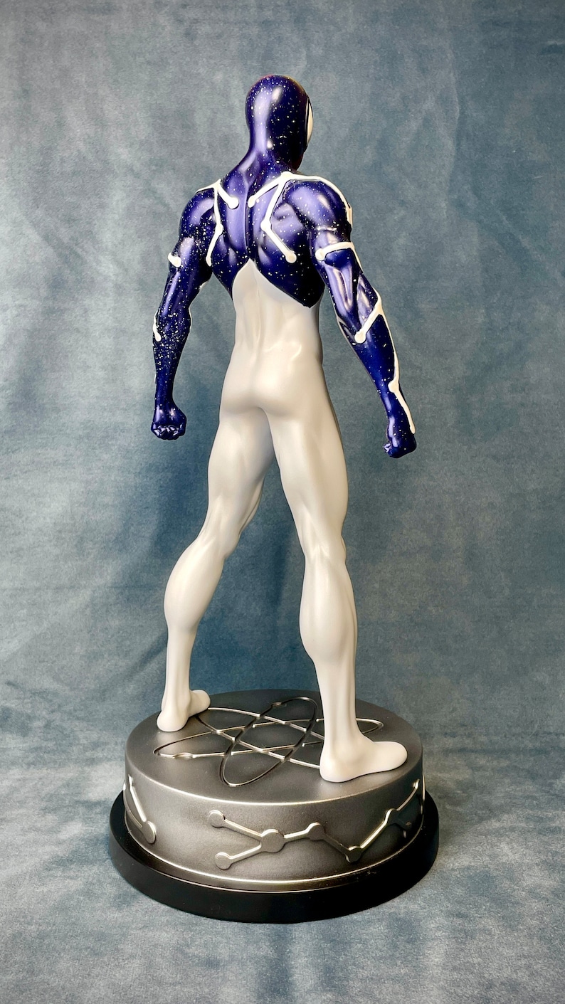 Puede incluir: Una figura de acci&oacute;n de un superh&eacute;roe de color blanco y azul de pie sobre una base plateada con un dise&ntilde;o de s&iacute;mbolo at&oacute;mico. La figura tiene un cuerpo blanco con detalles azules y estrellas blancas en las &aacute;reas azules.