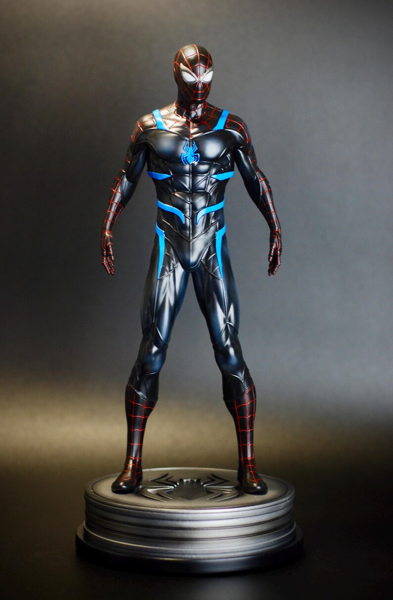 May include: A black and blue superhero figurine with red accents, standing on a circular base. The figure has a detailed costume with a spider emblem on the chest and a web-like pattern on the head and limbs. The figurine is approximately 12 inches tall.