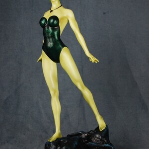 May include: A green-skinned figurine of a woman wearing a dark green swimsuit. The figurine is standing on a black rock base.