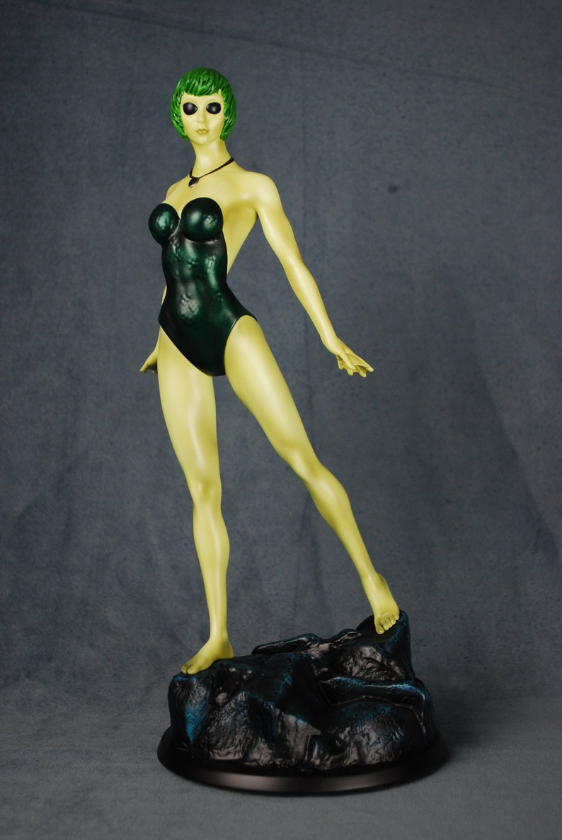 May include: A green-skinned figurine of a woman wearing a dark green swimsuit. The figurine is standing on a black rock base.