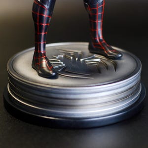 May include: A black and red superhero figure's legs and feet stand on a silver, circular base. The base features a spider emblem and is layered with a black base. The figure's costume has a red web pattern.