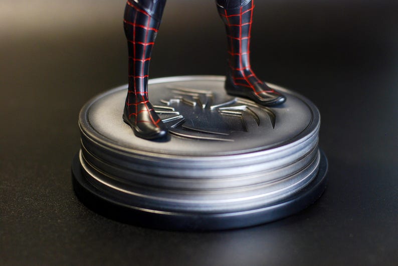 May include: A black and red superhero figure's legs and feet stand on a silver, circular base. The base features a spider emblem and is layered with a black base. The figure's costume has a red web pattern.