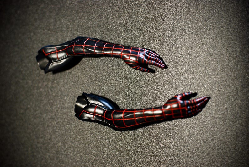 May include: Two black and red action figure arms, featuring a webbed pattern. The arms are posed with open hands, showcasing the intricate design and detail. The figure arms are set against a dark gray background.