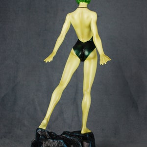 May include: A green-haired figurine of a person wearing a dark green swimsuit. The figurine is standing on a black base with a rocky texture.