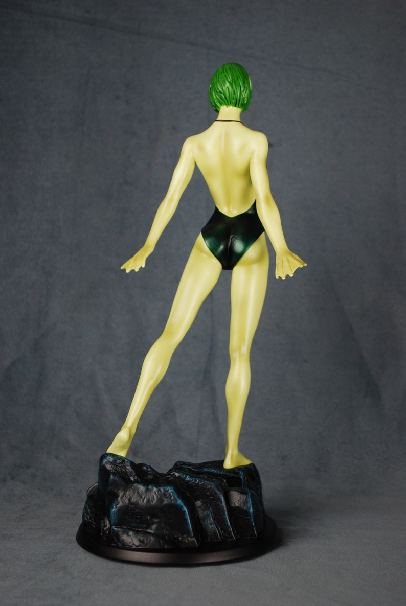 May include: A green-haired figurine of a person wearing a dark green swimsuit. The figurine is standing on a black base with a rocky texture.