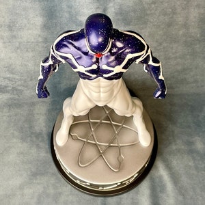Puede incluir: Una figura de acci&oacute;n blanca y azul de un superh&eacute;roe de pie sobre una base redonda con un s&iacute;mbolo de &aacute;tomo plateado. La figura tiene un cuerpo azul oscuro con l&iacute;neas blancas y una gema roja en el pecho.