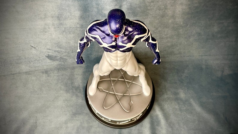 Puede incluir: Una figura de acci&oacute;n blanca y azul de un superh&eacute;roe de pie sobre una base redonda con un s&iacute;mbolo de &aacute;tomo plateado. La figura tiene un cuerpo azul oscuro con l&iacute;neas blancas y una gema roja en el pecho.