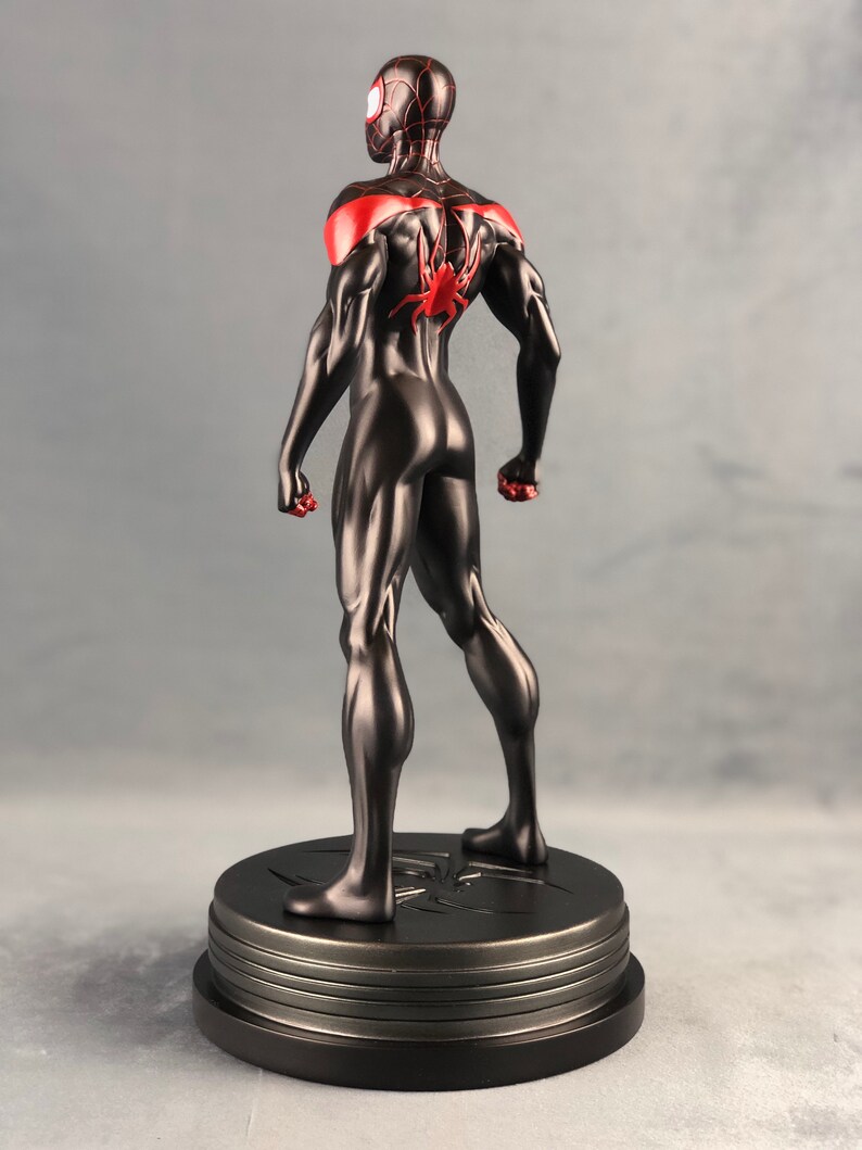 May include: Black and red superhero action figure standing on a black base with a spider symbol. The figure is posed with arms outstretched and legs slightly apart.