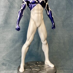 Puede incluir: Una figura de acci&oacute;n blanca y morada de un superh&eacute;roe que lleva una m&aacute;scara y un traje con un patr&oacute;n de estrellas. La figura est&aacute; de pie sobre una base plateada con un s&iacute;mbolo de &aacute;tomo.