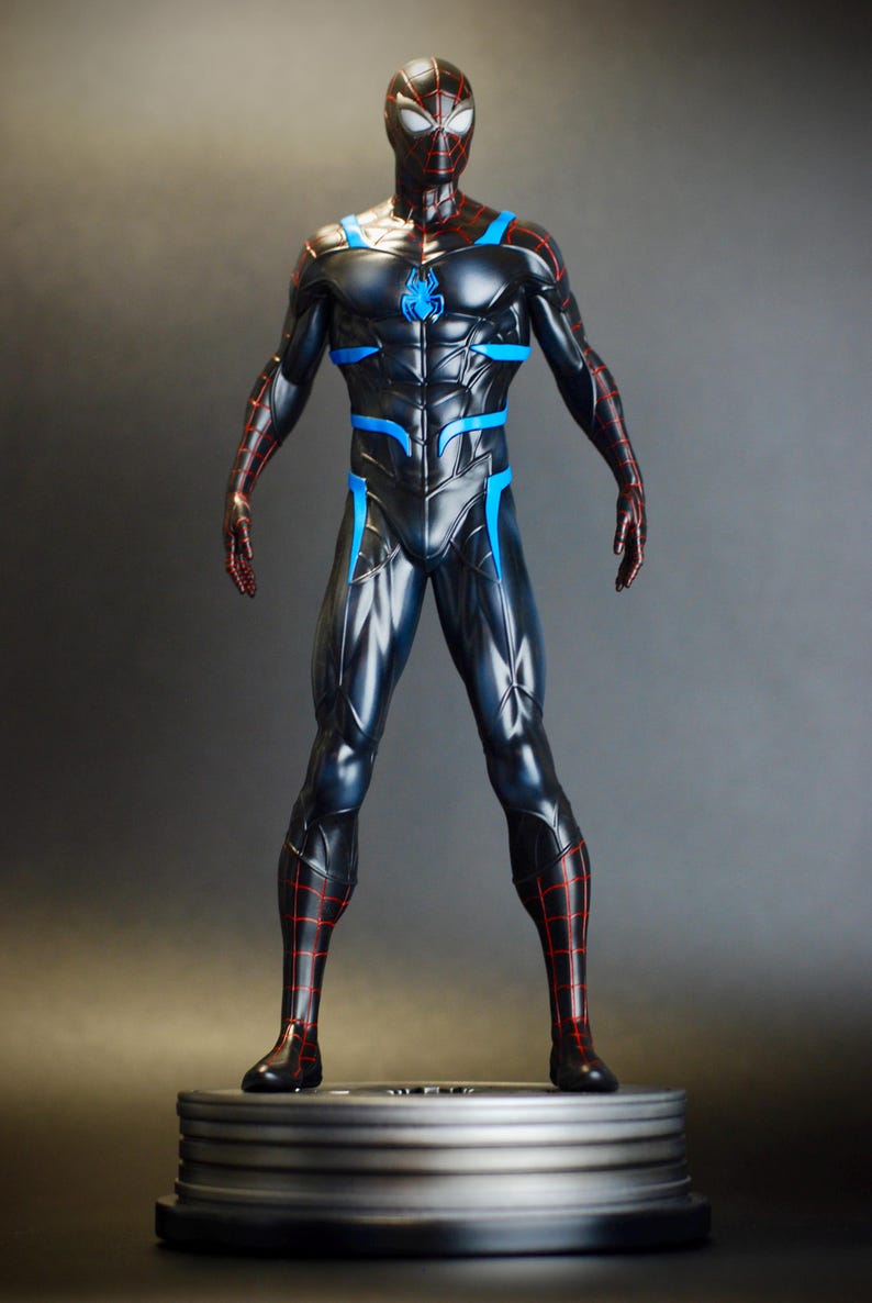 May include: A black and blue superhero figurine with red accents. The figure is standing on a round, metallic base. The superhero suit features a spider emblem on the chest and blue highlights. The figure's mask has a unique design.