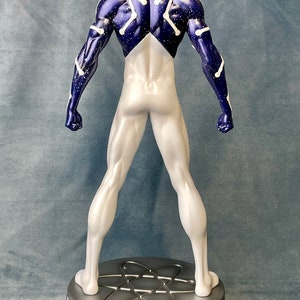 Puede incluir: Una figura de superh&eacute;roe blanca y azul con un patr&oacute;n de constelaci&oacute;n en la espalda. La figura est&aacute; de pie sobre una base plateada con un dise&ntilde;o de &aacute;tomo estilizado.