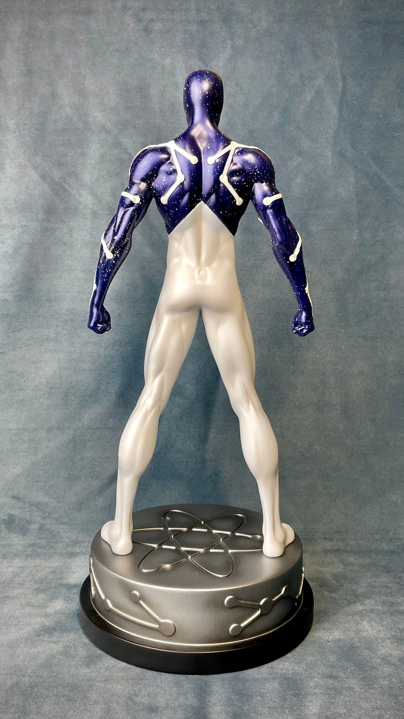 Puede incluir: Una figura de superh&eacute;roe blanca y azul con un patr&oacute;n de constelaci&oacute;n en la espalda. La figura est&aacute; de pie sobre una base plateada con un dise&ntilde;o de &aacute;tomo estilizado.