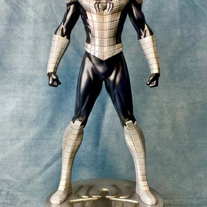 May include: A silver and black action figure of Spider-Man standing on a silver base with a spider logo. The figure is wearing a silver and black suit with a black mask.