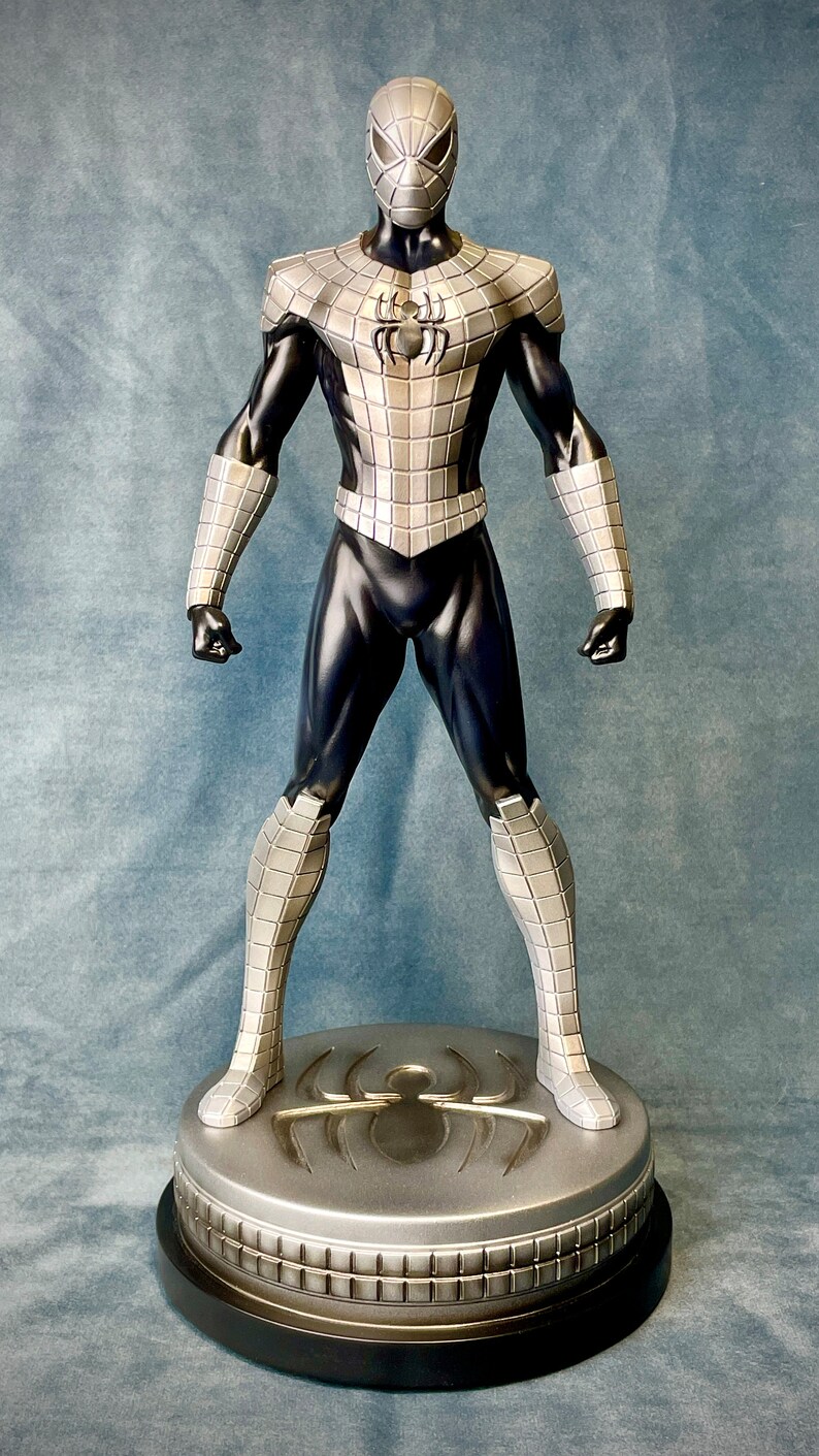 May include: A silver and black action figure of Spider-Man standing on a silver base with a spider logo. The figure is wearing a silver and black suit with a black mask.