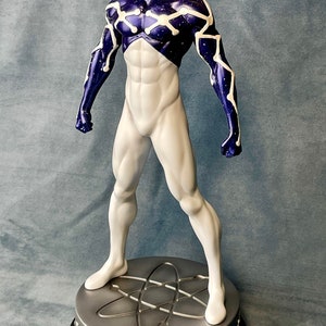 Puede incluir: Una figura de acci&oacute;n blanca y azul de un superh&eacute;roe con una m&aacute;scara roja en forma de ara&ntilde;a. La figura est&aacute; de pie sobre una base plateada con un dise&ntilde;o de s&iacute;mbolo at&oacute;mico.