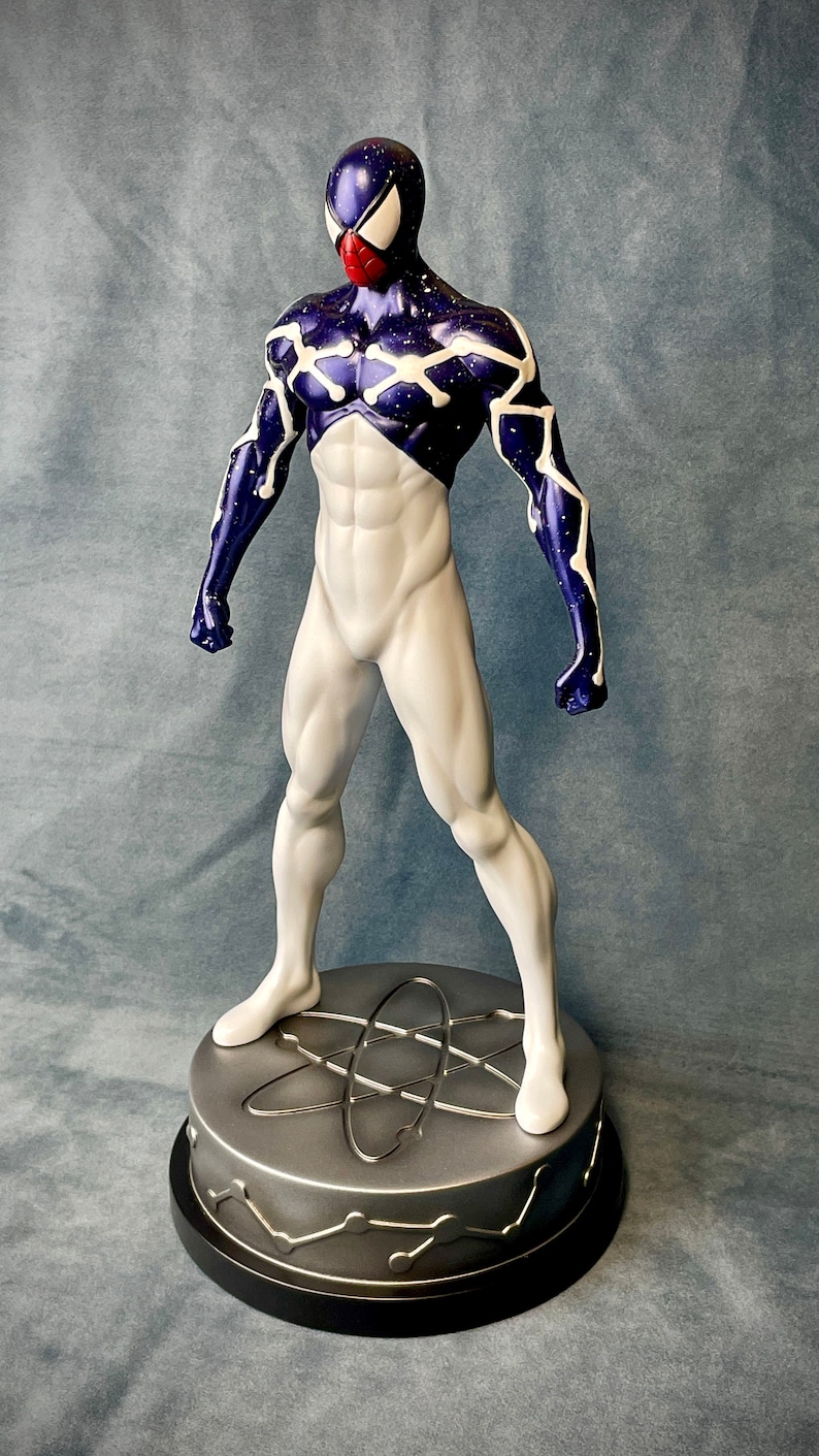 Puede incluir: Una figura de acci&oacute;n blanca y azul de un superh&eacute;roe con una m&aacute;scara roja en forma de ara&ntilde;a. La figura est&aacute; de pie sobre una base plateada con un dise&ntilde;o de s&iacute;mbolo at&oacute;mico.