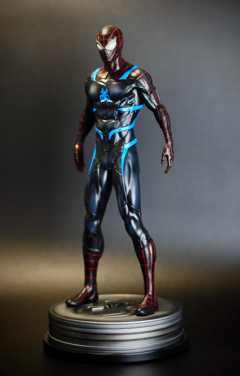 May include: A black and blue superhero figurine with red accents, standing on a silver base. The figure has a detailed costume with a spiderweb pattern and blue highlights. The figurine is a collectible item.