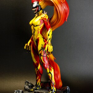 May include: A detailed figurine of a female character with a fiery red and yellow color scheme. The figure has a black and white face, and the hair flows dramatically. The character is posed on a rocky base, with a black background.