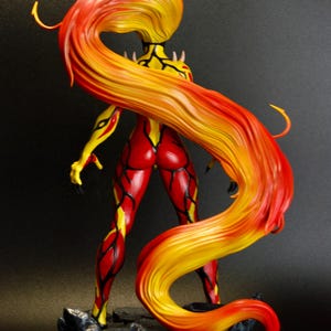 May include: A figurine of a female character with fiery red and yellow hair cascading down her back. The figure is in a dynamic pose, with red and yellow detailing on the body. The base is a dark, textured surface.