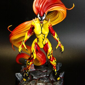 May include: A detailed figurine of a female superhero with a yellow and red color scheme. The figure has long, flowing orange and red hair. The base is a rocky terrain with a black circular base. The figure is in a dynamic pose, with sharp claws and a menacing expression.