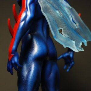 May include: A blue and red action figure with a translucent blue cape. The figure is posed with its back to the camera, showing off its muscular physique.