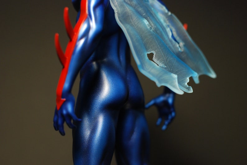 May include: A blue and red action figure with a translucent blue cape. The figure is posed with its back to the camera, showing off its muscular physique.