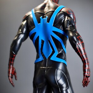 May include: A black action figure with a blue spider emblem on the back. The figure has red lines on the arms and hands. The figure is a superhero.