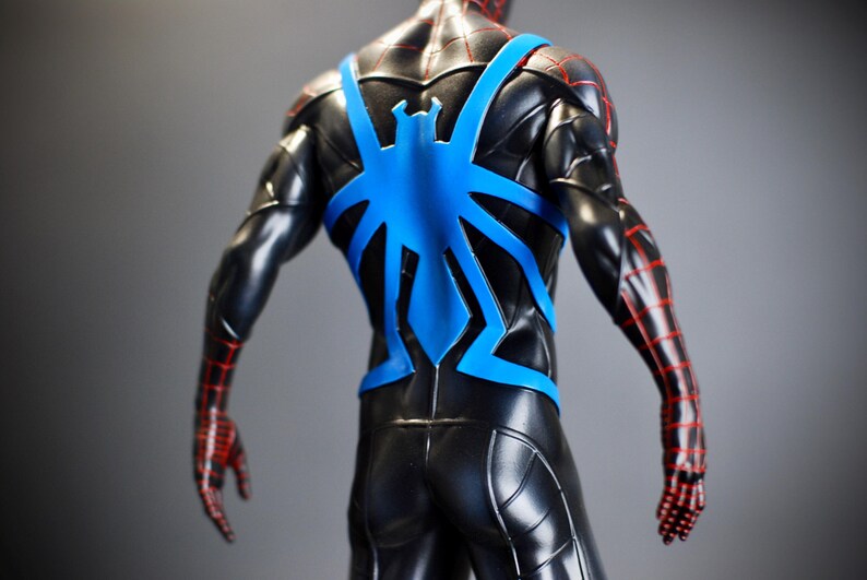 May include: A black action figure with a blue spider emblem on the back. The figure has red lines on the arms and hands. The figure is a superhero.