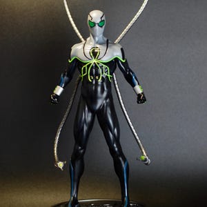 Puede incluir: Una figura de acci&oacute;n negra y verde de un superh&eacute;roe con cuatro brazos mec&aacute;nicos. La figura est&aacute; de pie sobre una base plateada con un dise&ntilde;o circular.