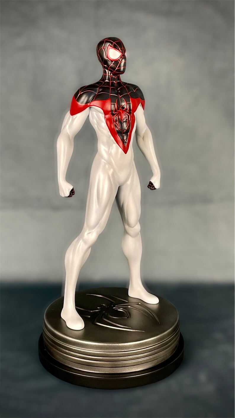 May include: A white and red action figure of Spider-Man with a black spider symbol on his chest. The figure is standing on a round, metallic base with a spider symbol.