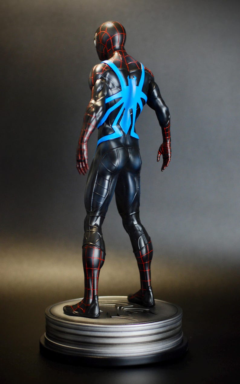 May include: A black and red superhero figure with a blue spider emblem on the back. The figure is standing on a silver circular base. The suit has red web-like detailing and the figure is posed with its back to the viewer.