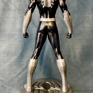 May include: A black and silver action figure of Spider-Man standing on a circular base with a spider logo. The figure is posed with its arms at its sides and its legs slightly apart.
