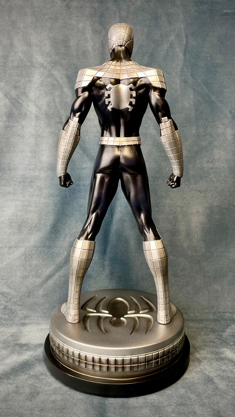 May include: A black and silver action figure of Spider-Man standing on a circular base with a spider logo. The figure is posed with its arms at its sides and its legs slightly apart.